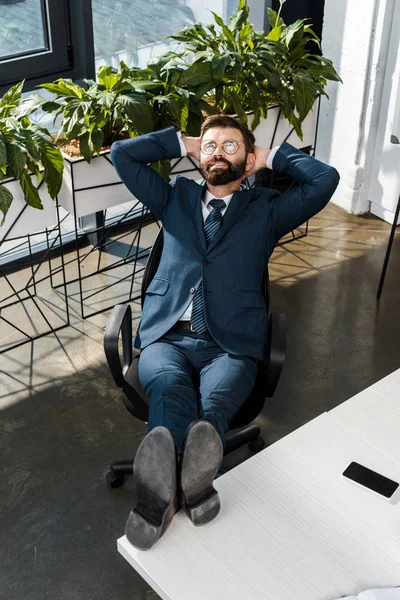 Vue grand angle d'homme d'affaires barbu souriant relaxant avec les jambes sur la table et les mains derrière la tête au bureau — Photo de stock