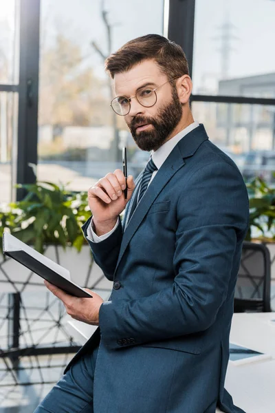 Serio hombre de negocios barbudo sosteniendo bloc de notas y mirando a la cámara en la oficina — Stock Photo