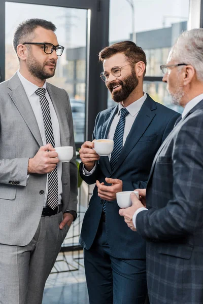 Tres hombres de negocios sonrientes en ropa formal bebiendo café y hablando en la oficina - foto de stock