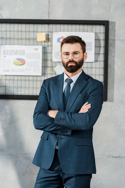 Homem de negócios barbudo confiante em terno e óculos de pé com braços cruzados e olhando para a câmera no escritório — Fotografia de Stock