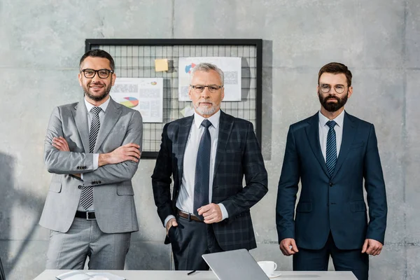 Drei selbstbewusste Geschäftsleute, die im Büro zusammenstehen und in die Kamera schauen — Stockfoto