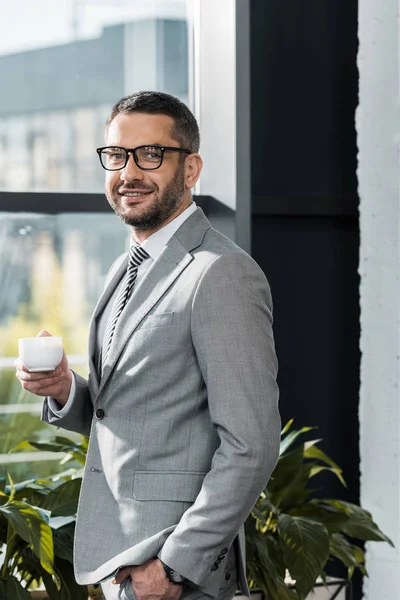 Guapo feliz hombre de negocios sosteniendo taza de café y sonriendo a la cámara en la oficina - foto de stock