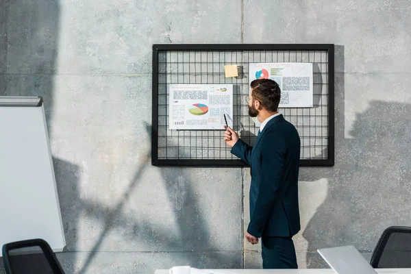 Seitenansicht eines bärtigen Geschäftsmannes, der Stift in der Hand hält und auf Geschäftstabellen an der Wand zeigt — Stockfoto