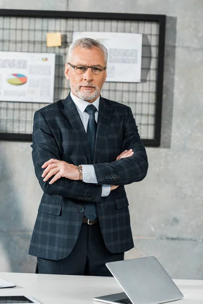 Selbstbewusster reifer Geschäftsmann, der mit verschränkten Armen im Büro steht und in die Kamera blickt — Stockfoto