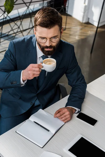 Hochwinkelaufnahme des bärtigen Geschäftsmannes in Brille, der am Arbeitsplatz Kaffee trinkt — Stockfoto