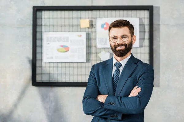 Красивий впевнений бородатий бізнесмен стоїть з схрещеними руками і посміхається на камеру в офісі — стокове фото