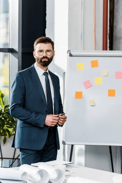 Ernster bärtiger Geschäftsmann blickt in die Kamera, während er Stift in der Hand hält und neben Whiteboard steht — Stockfoto