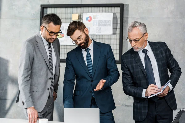 Tres hombres de negocios profesionales serios mirando el ordenador portátil y discutiendo el proyecto en la oficina - foto de stock