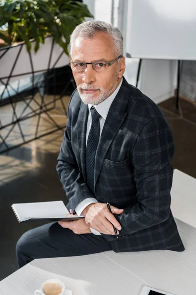 Hochwinkelaufnahme eines ernsthaften reifen Geschäftsmannes, der auf dem Bürotisch sitzt und in die Kamera schaut — Stockfoto