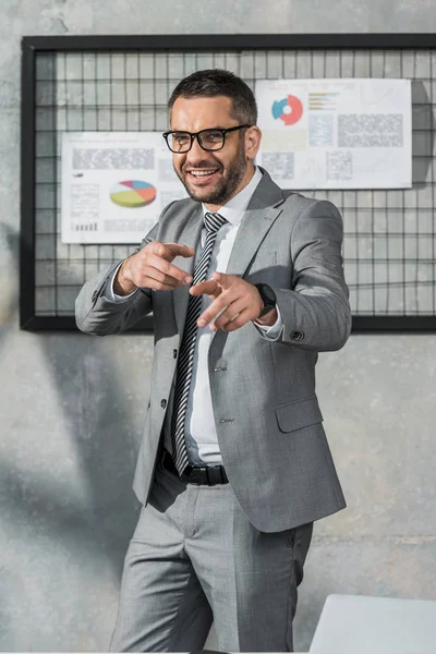 Cheerful businessman in suit and eyeglasses pointing with fingers and smiling at camera in office — Stock Photo