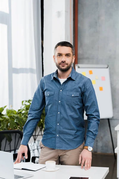 Handsome bearded businessman standing at workplace and looking at camera — Stock Photo