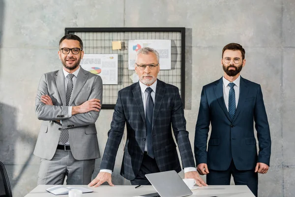 Drei selbstbewusste Geschäftsleute in formaler Kleidung, die im Büro zusammenstehen und in die Kamera schauen — Stockfoto