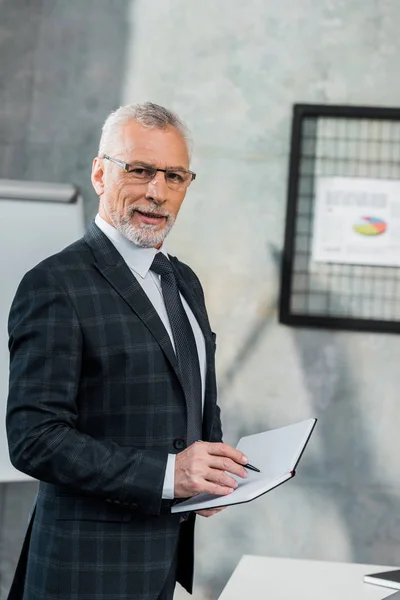 Mature businessman holding notebook and smiling at camera in office — Stock Photo