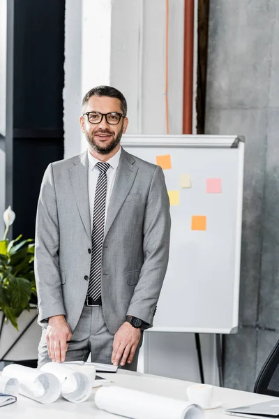 Bell'uomo d'affari in giacca e cravatta e occhiali in piedi sul posto di lavoro e sorridente alla fotocamera — Foto stock