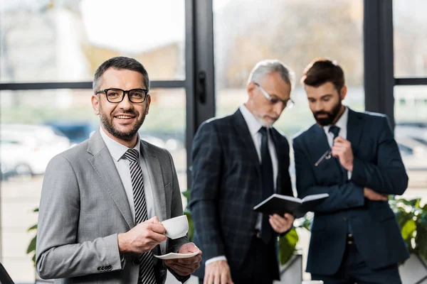 Handsome businessman drinking coffee and smiling at camera while colleagues working behind — Stock Photo