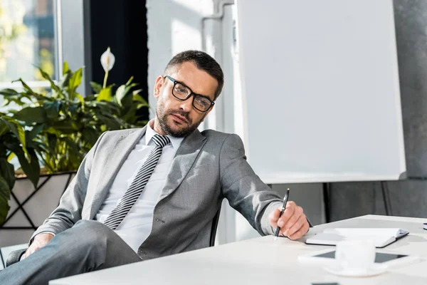 Скучающий бизнесмен в костюме и очках сидит и смотрит в сторону в офисе — стоковое фото