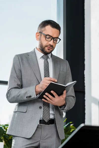 Bel homme d'affaires barbu en costume et lunettes debout et écrit dans un cahier sur le lieu de travail — Photo de stock