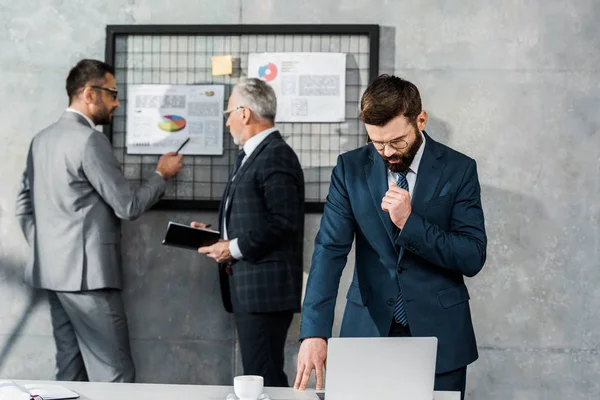 Hombre de negocios barbudo utilizando el ordenador portátil y mirando hacia abajo, mientras que los colegas que trabajan con gráficos de negocios detrás - foto de stock