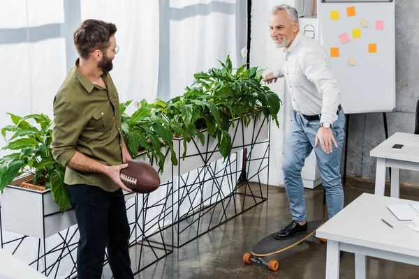 Uomini d'affari sorridenti divertirsi con skateboard e palla da rugby in ufficio — Foto stock