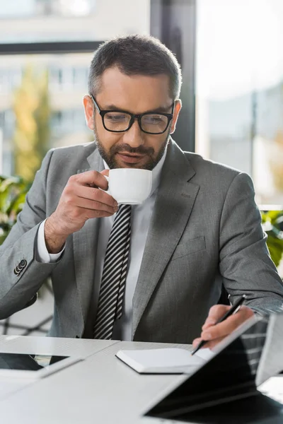 Businessman in suit and eyeglasses drinking coffee and writing in notebook at wokrplace — Stock Photo
