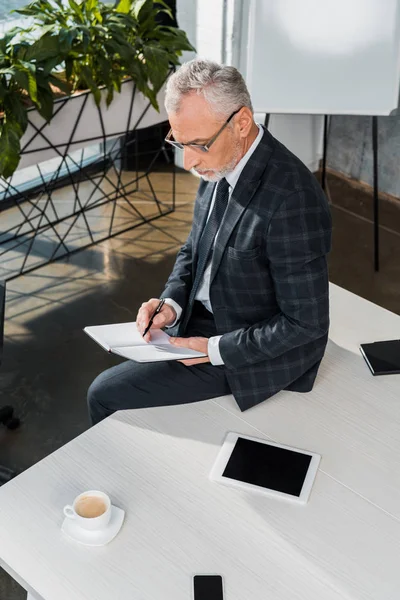 High angle view of mature businessman writing in notebook while sitting on office table — Stock Photo