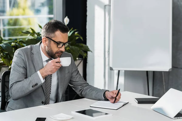 Целенаправленный бизнесмен в очках пьет кофе и пишет в блокноте на рабочем месте — стоковое фото