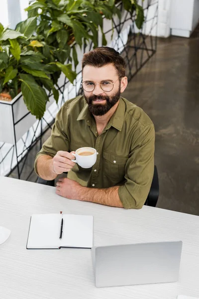 High angle view of bearded businessman in eyeglasses holding cup of coffee and smiling at camera while sitting at workplace — Stock Photo