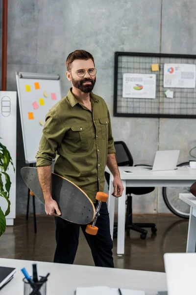 Schöner bärtiger Geschäftsmann mit Brille, Skateboard in der Hand und lächelnd in die Kamera im Büro — Stockfoto
