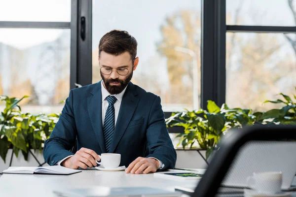 Sérieux homme d'affaires barbu en costume et lunettes tenant tasse de café tout en étant assis sur le lieu de travail — Photo de stock