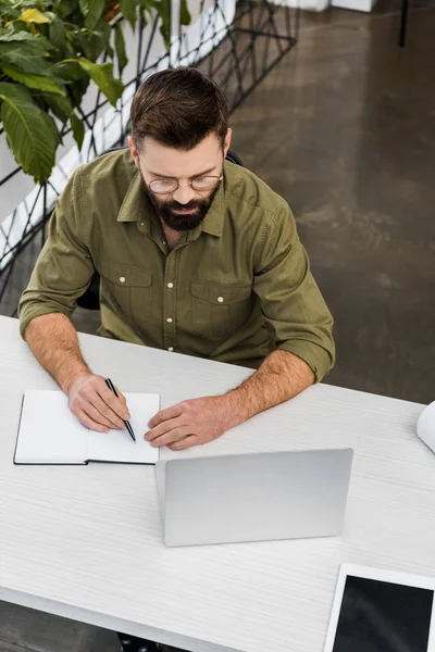Vista de ángulo alto de hombre de negocios barbudo en gafas de escribir en cuaderno y el uso de ordenador portátil en el lugar de trabajo - foto de stock