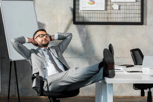 Relaxed buisnessman in eyeglasses sitting with legs on table and hands behind head in office — Stock Photo