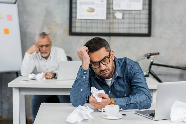 Bored buisnessman in eyeglasses holding crumpled paper while sitting at workplace — Stock Photo