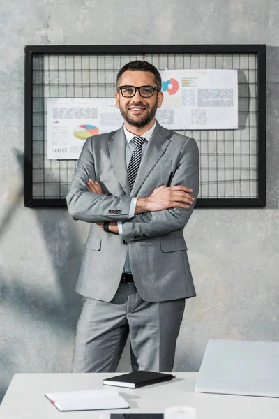 Handsome businessman in suit and eyeglasses standing with crossed arms and smiling at camera in office — Stock Photo