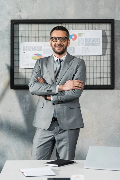 Bel homme d'affaires en costume et lunettes debout avec les bras croisés et souriant à la caméra dans le bureau — Photo de stock