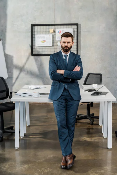 Vista completa di uomo d'affari barbuto fiducioso in giacca e occhiali in piedi con braccia incrociate in ufficio — Foto stock