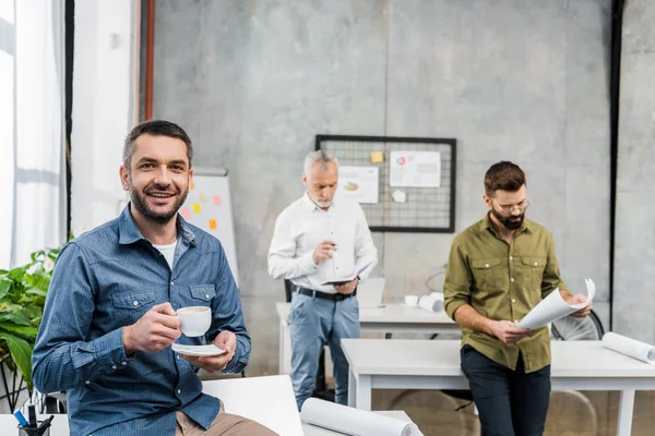 Schöner Geschäftsmann mit einer Tasse Kaffee, der in die Kamera lächelt, während Kollegen hinter ihm aufwachen — Stockfoto