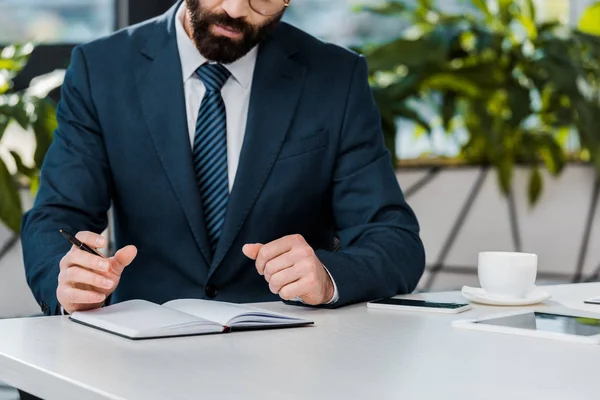 Recortado tiro de barbudo empresario celebración de pluma y escritura en cuaderno - foto de stock