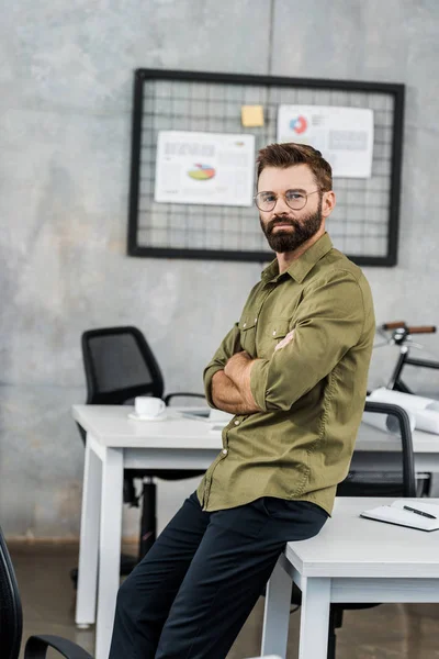 Schöner bärtiger Geschäftsmann mit Brille sitzt mit verschränkten Armen und blickt in die Kamera im Büro — Stockfoto
