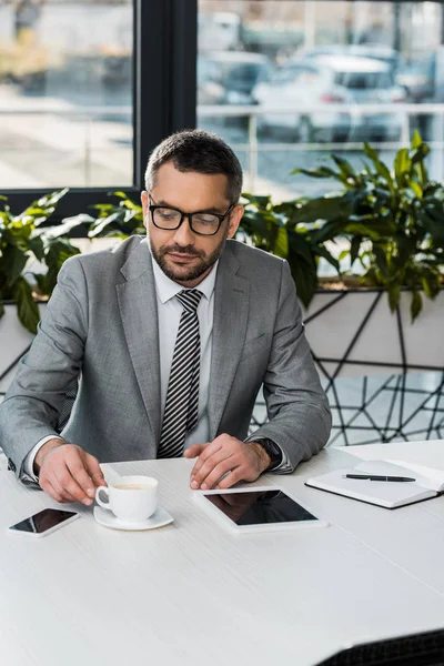 Hombre de negocios serio en traje y anteojos sentado en el lugar de trabajo y mirando una taza de café - foto de stock