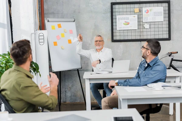 Smiling mature businessman throwing paper plane to colleagues in office — Stock Photo