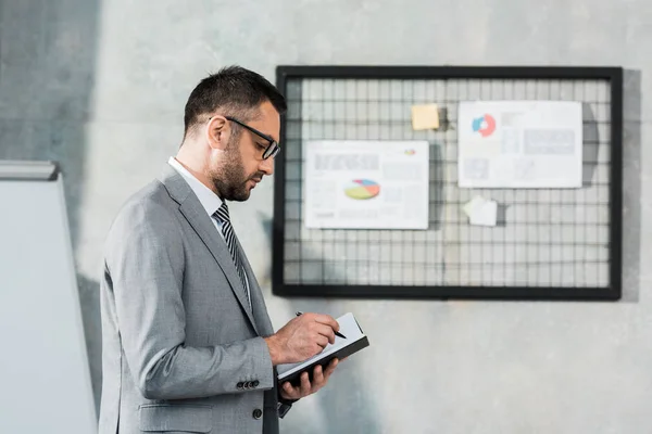 Seitenansicht eines Geschäftsmannes mit Brille, der am Arbeitsplatz Notizen im Notizblock macht — Stockfoto