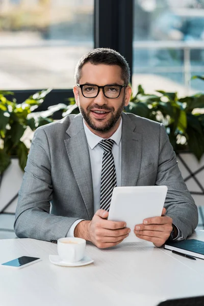 Cheerful handsome businessman holding tablet and looking at camera in office — Stock Photo