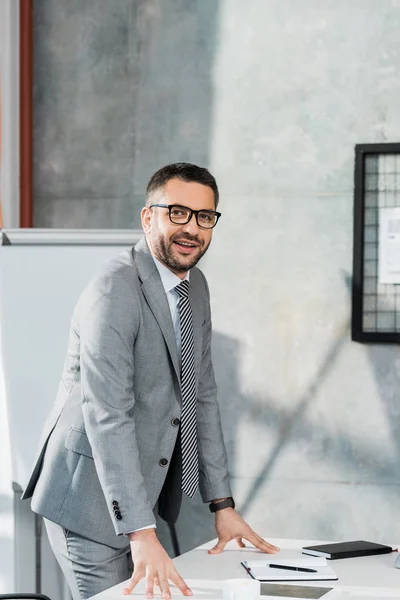 Lächelnder gutaussehender Geschäftsmann in Anzug und Brille, der sich auf den Tisch lehnt und im Büro in die Kamera schaut — Stockfoto