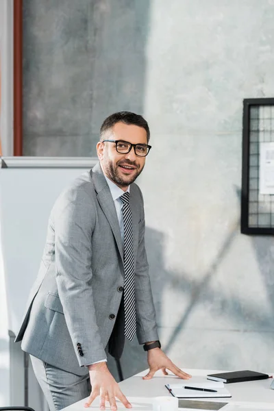 Beau homme d'affaires souriant en costume et lunettes appuyé sur la table et regardant la caméra dans le bureau — Photo de stock