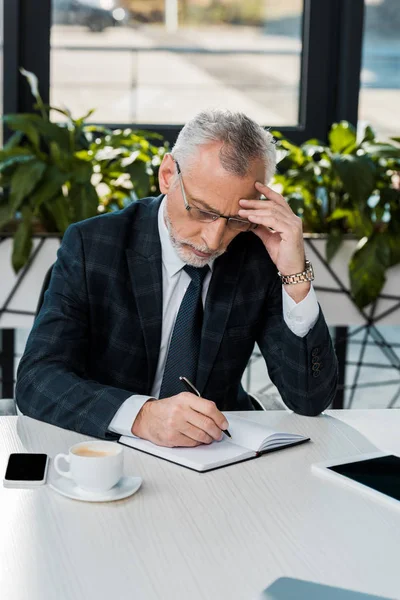 Nachdenklich gutaussehender Geschäftsmann mittleren Alters im Anzug schreibt im Büro etwas ans Notizbuch — Stockfoto