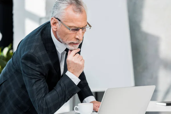 Pensativo guapo mediana edad hombre de negocios en traje y gafas mirando portátil en la oficina - foto de stock