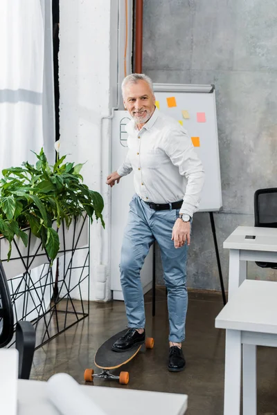 Cheerful handsome middle aged businessman putting leg on longboard in office — Stock Photo