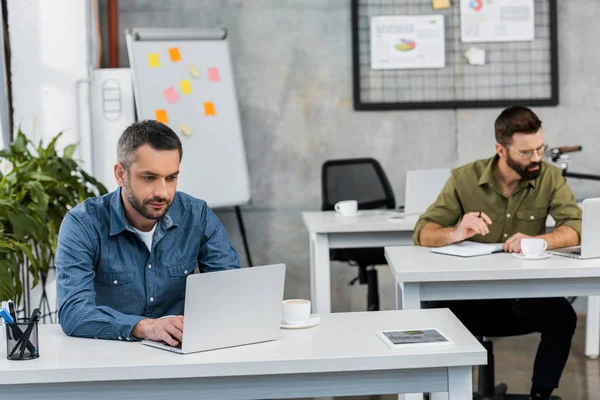 Dos hombres de negocios guapos que trabajan en computadoras portátiles en la oficina - foto de stock