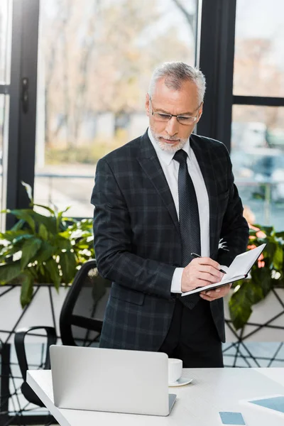 Gutaussehender Geschäftsmann mittleren Alters schreibt an Notizbuch und schaut im Büro auf Laptop — Stockfoto