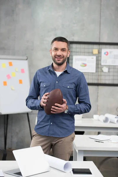 Sonriente hombre de negocios guapo sosteniendo pelota de fútbol americano y mirando a la cámara en la oficina - foto de stock